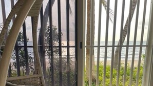 A room with a view of the beach through a barred window, a white bed with a grey blanket, and a white chair. - Studio Océan - Vue sur Mer/Pied dans l'eau (Libreville)