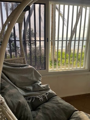A room with a view of the beach through a barred window, a white bed with a grey blanket, and a white chair.
