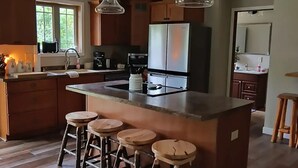 A modern kitchen with wooden cabinets, a central island with stools, pendant lighting, and a large refrigerator. - Troullier's River House

 (White Lake)
