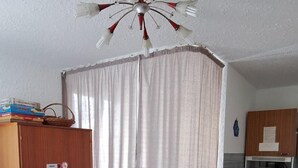 Una stanza con pavimento in legno, un tavolo da pranzo con tovaglia, un armadio, una tenda, una ventola a soffitto, un forno a microonde e un frigorifero.
