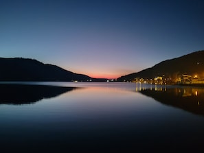 A calm lake at dusk with a sunset and a distant shoreline with buildings.