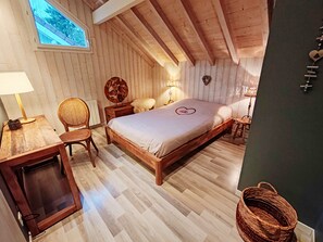 A bedroom with a wooden ceiling, a bed with a heart-shaped pillow, a desk with a lamp, and a window with a view of trees.