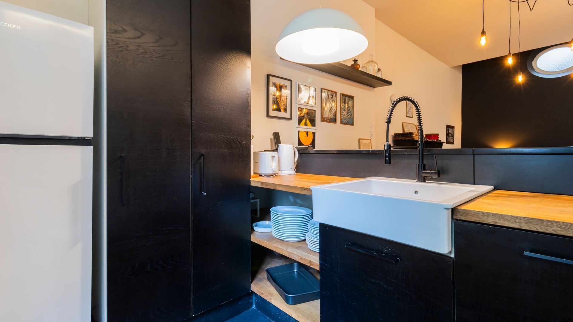Une cuisine moderne avec des meubles noirs, un évier blanc et un plan de travail en bois. Des tableaux encadrés ornent le mur, et une suspension lumineuse est placée au-dessus de l’évier.