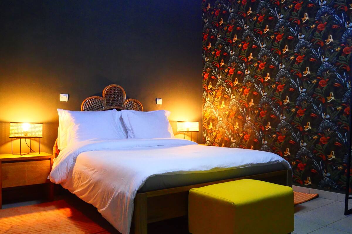 A neatly made bed with white linens, two bedside lamps, a wicker headboard, and a floral-patterned wall.
