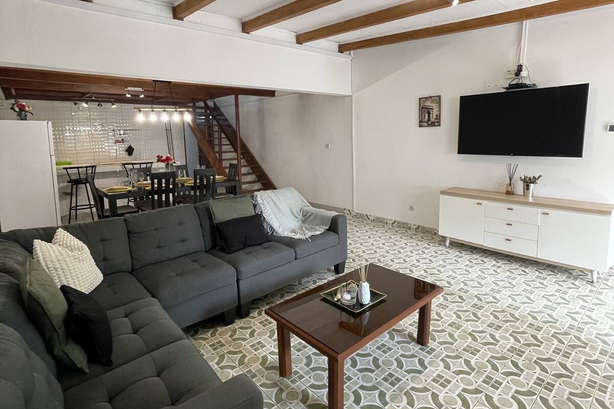 A spacious living area with a grey sectional sofa, a wooden coffee table, and a white cabinet with a flat-screen TV.