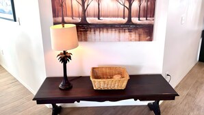 Una mesa de madera oscura con una lámpara y una cesta de mimbre, frente a una pared con un cuadro de árboles y un cuerpo de agua.