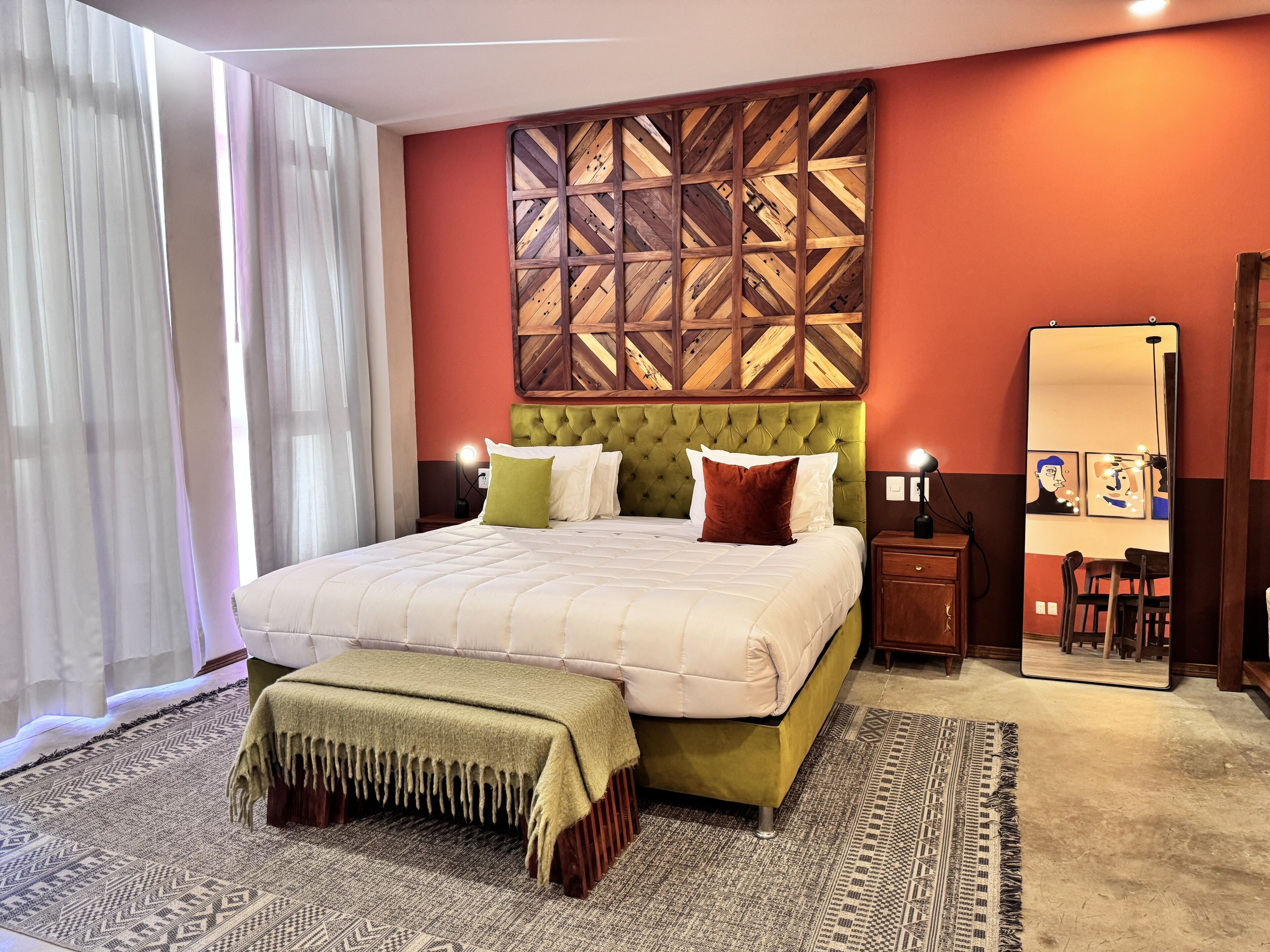 A hotel room with a large bed, a green upholstered headboard, a bedside table with a lamp, a mirror, and a patterned rug.