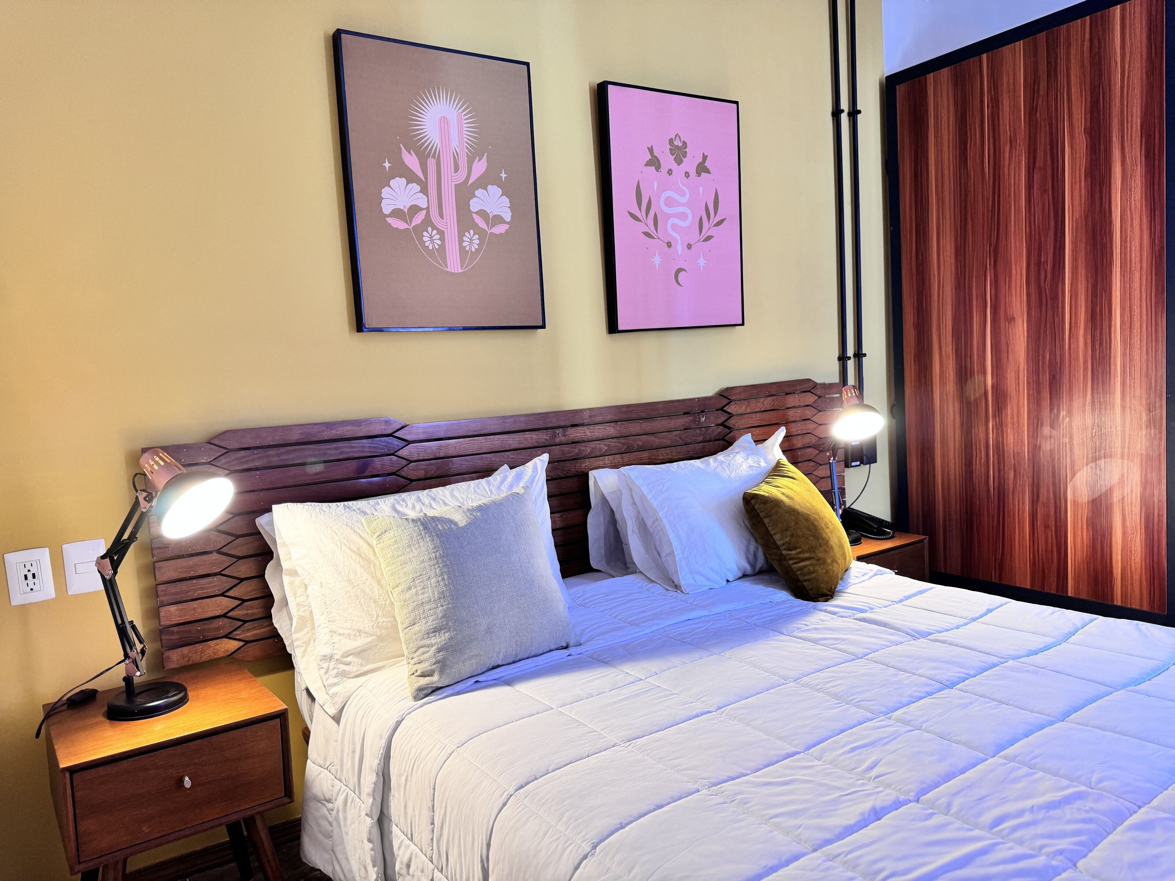 A neatly made bed with white bedding and multiple pillows, a wooden headboard, bedside lamps, and two framed artworks on the wall.
