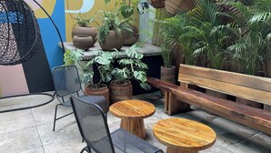 Un área de estar al aire libre con sillas de metal y mesas de madera, rodeada de plantas en macetas y un mural.