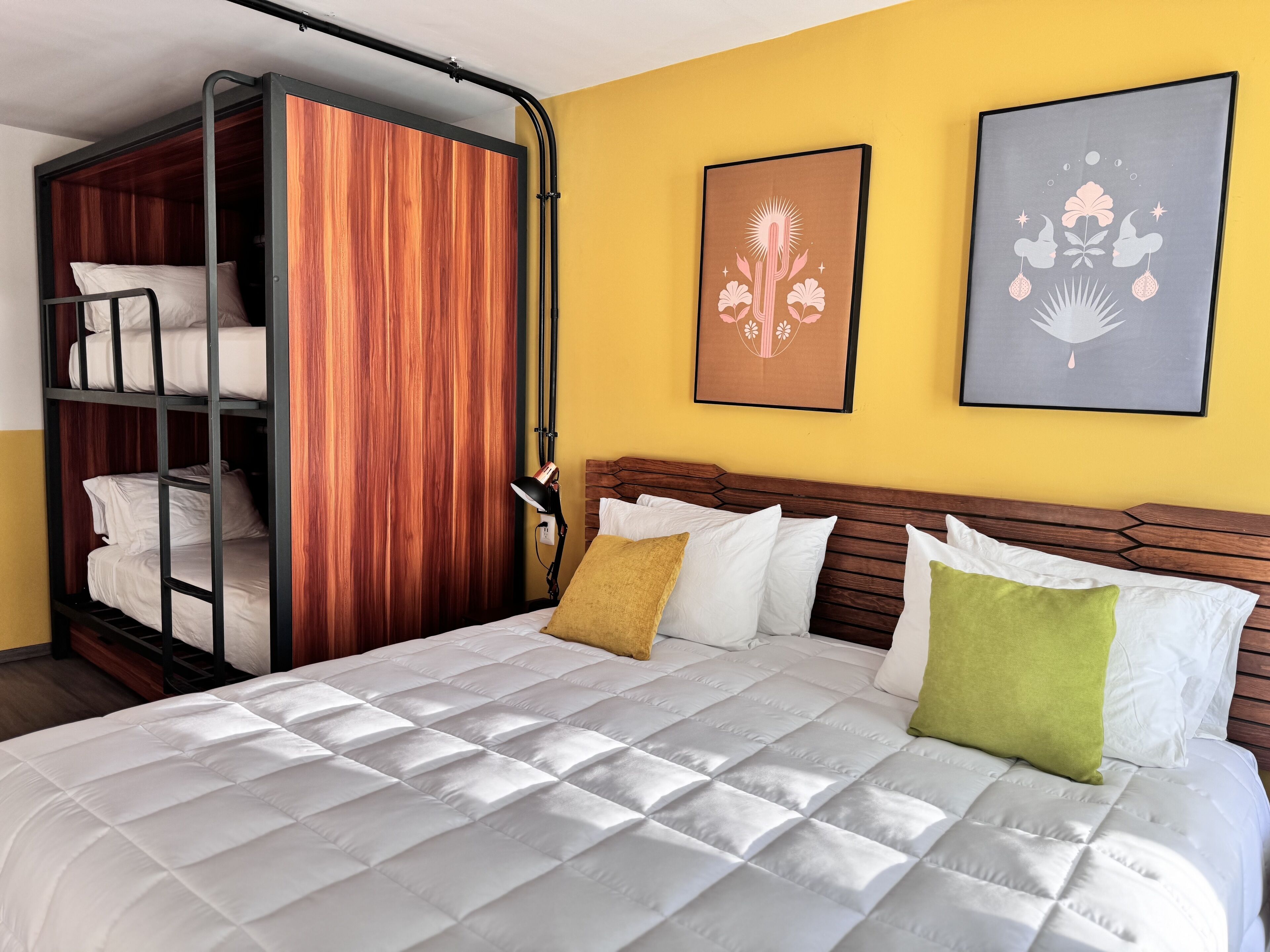 A hotel room with a bunk bed, a wooden wardrobe, a bed with white bedding and pillows, and two framed artworks on the wall.