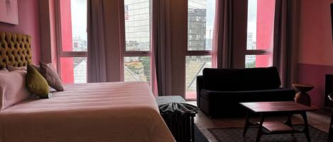 Habitación de hotel con una cama grande, un sofá negro, una mesita y vistas a la ciudad a través de amplios ventanales.