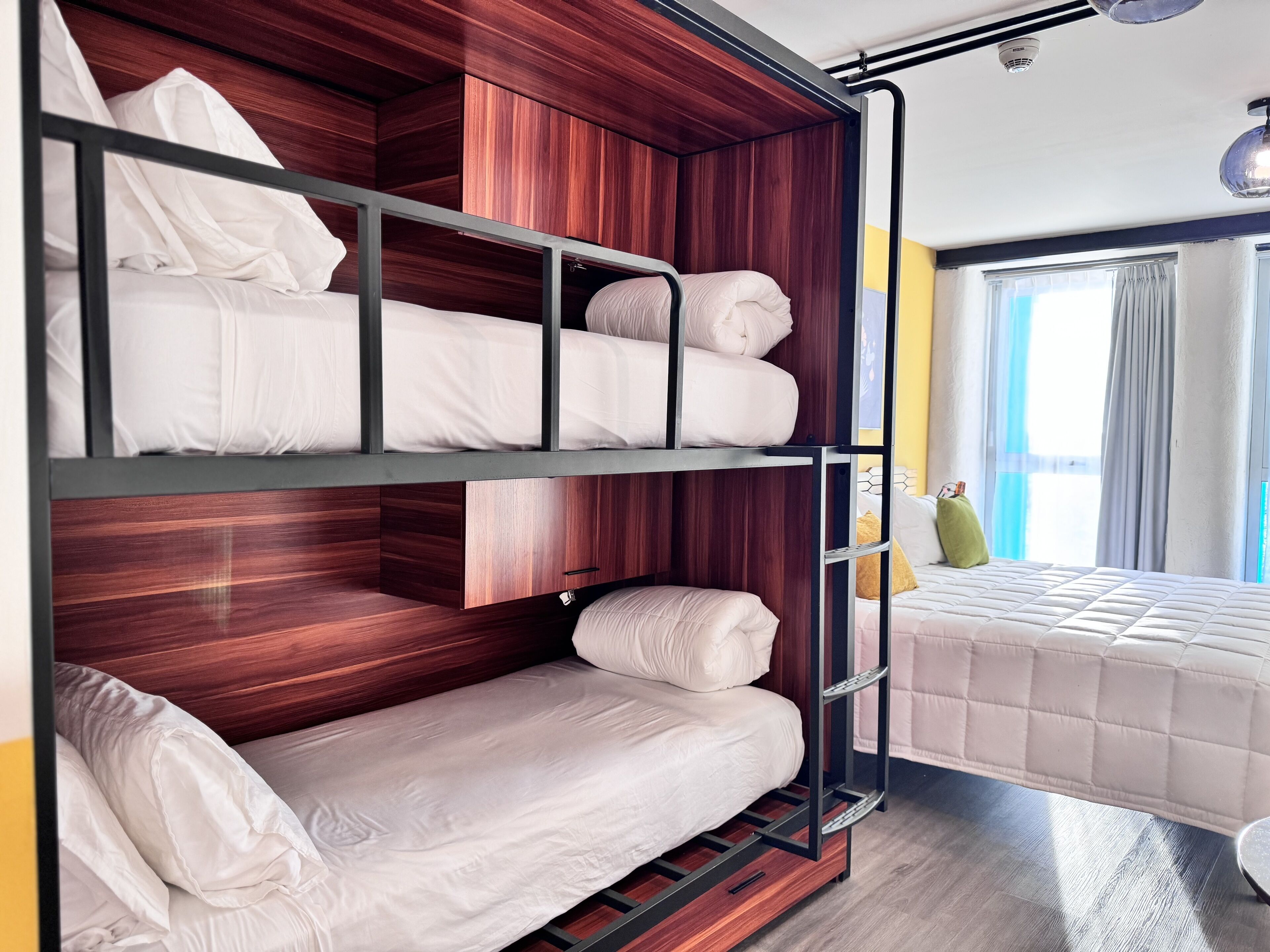 A bunk bed with wooden panels and metal railings, a window with curtains, and a bed with a quilted cover.