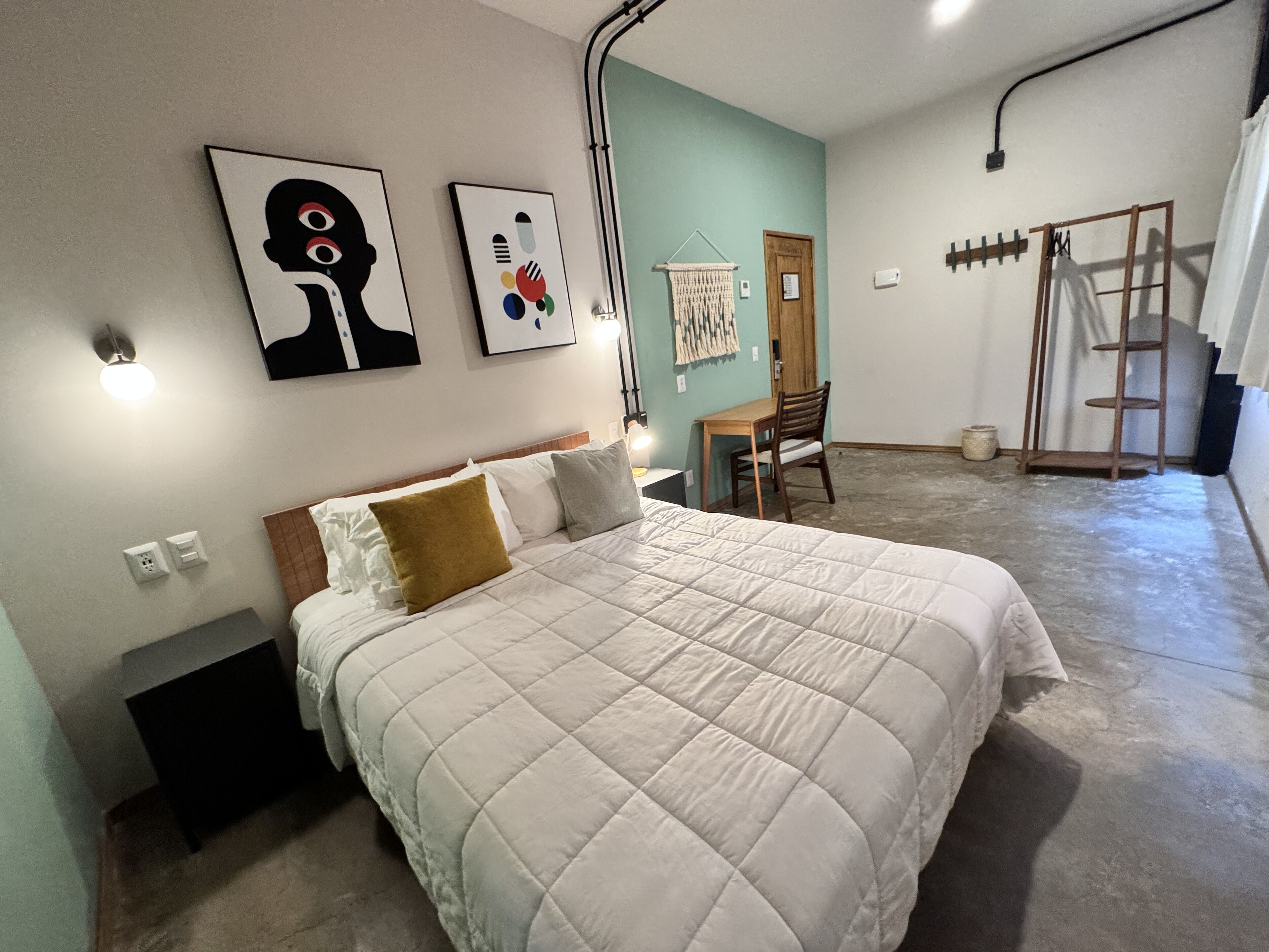 Un dormitorio con una cama grande, un escritorio y una silla. Hay obras de arte en las paredes y una puerta.