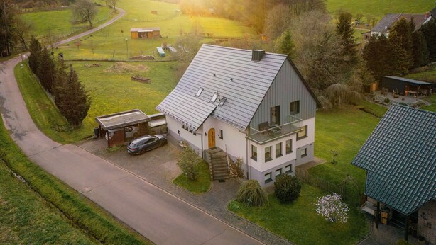 A two-story house with a sloped roof, a car parked in front, and a small garden with a flower bed. - Beautiful, spacious holiday apartment in Neroth (Neroth)