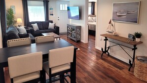 A modern living room with a dark wood floor, a black sofa with patterned cushions, a dining table with chairs, a TV mounted on the wall, and a painting of boats. - Land Lovers & Pirates Welcome #3 (Port St. Joe)