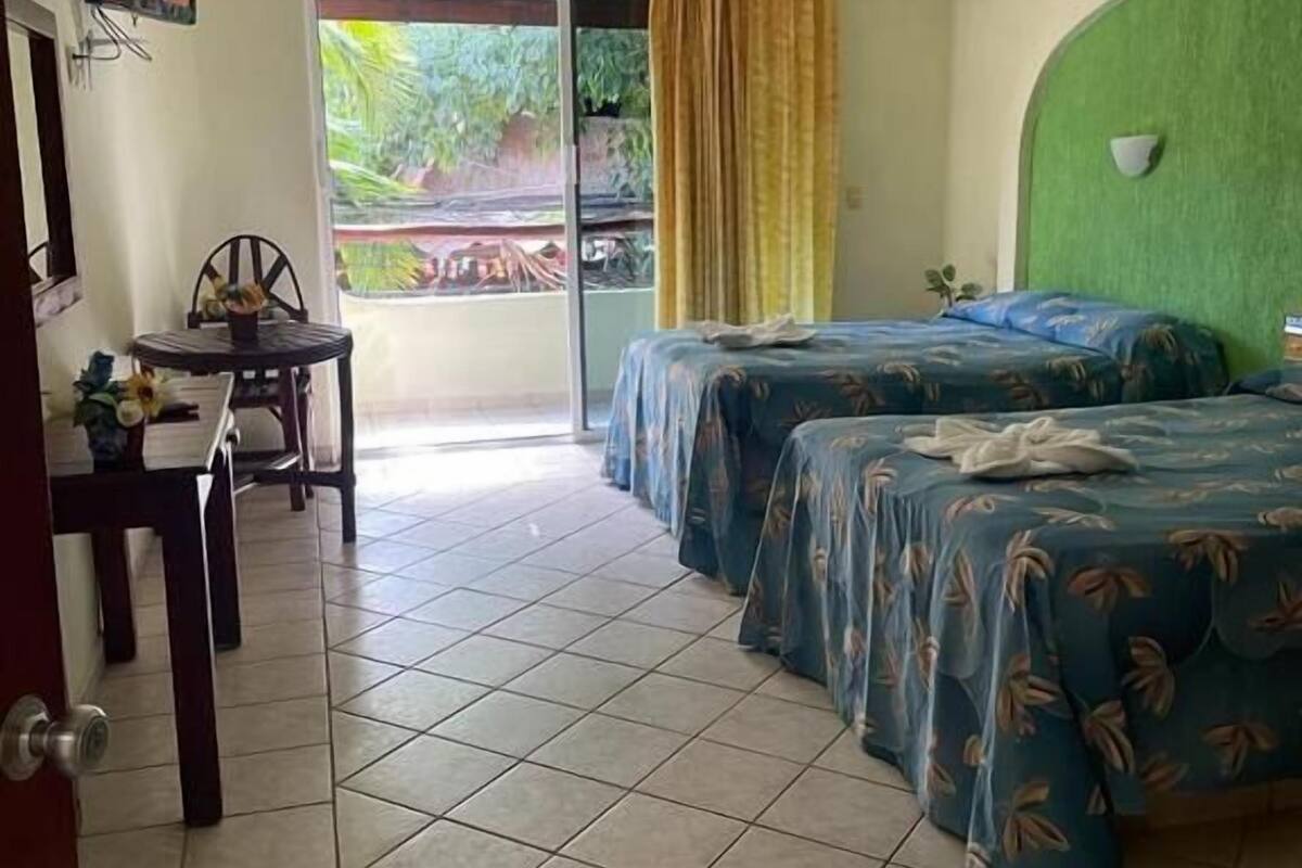 Habitación de hotel con dos camas, un escritorio, una silla, ventilador de techo y vistas al exterior.