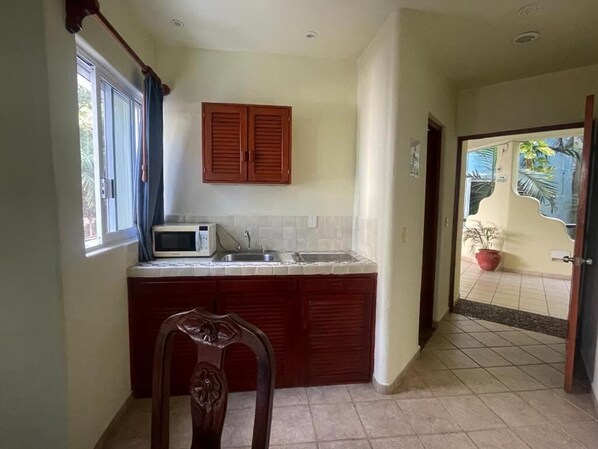 A compact kitchen with a microwave, sink, and wooden cabinets. A chair is placed in front of the counter. - HOTEL CASTILLO DEL MAR (Playa del Carmen)