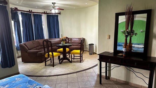 A hotel room with a bed, a dining area with a brown sofa and yellow chairs, a TV on a stand, and a ceiling fan.