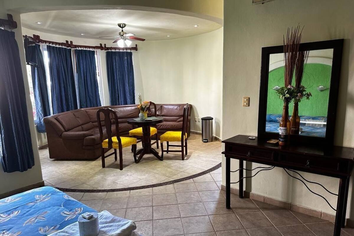 Una habitación de hotel con cama, zona de comedor con un sofá marrón y sillas amarillas, televisor en un pedestal y ventilador de techo.