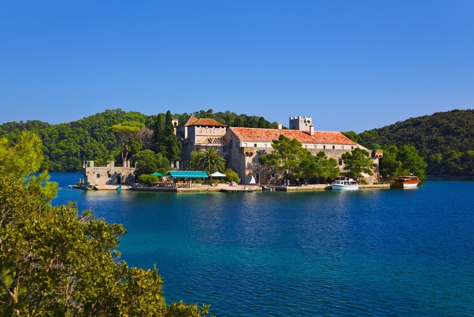 A historic building with a tower, situated on a lakeside with clear blue water and lush greenery.