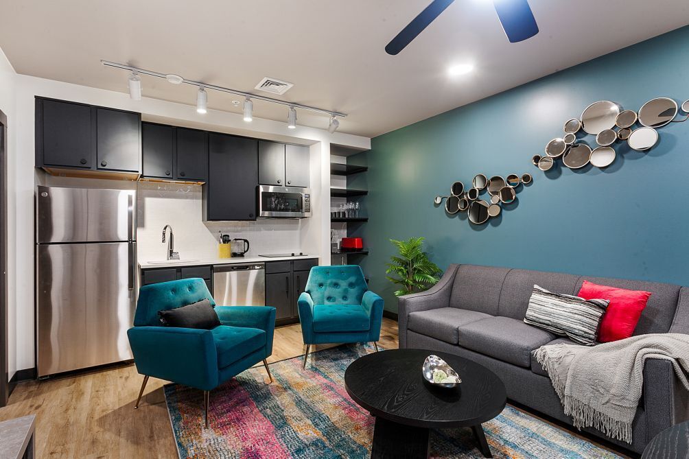 A modern kitchen with stainless steel appliances, teal armchairs, a gray sofa, and a colorful rug.