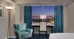 Ein Hotelzimmer mit Bett, Schreibtisch mit Stuhl, Blick auf die Stadtlandschaft und einer Stehlampe.