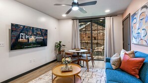 Una sala de estar moderna con una televisión mostrando 'Hosteva Hotel Standards, Comfort of a Home', un ventilador de techo, un comedor con mesa y sillas, un sofá azul y un amplio ventanal con vista a un muro de ladrillo.
