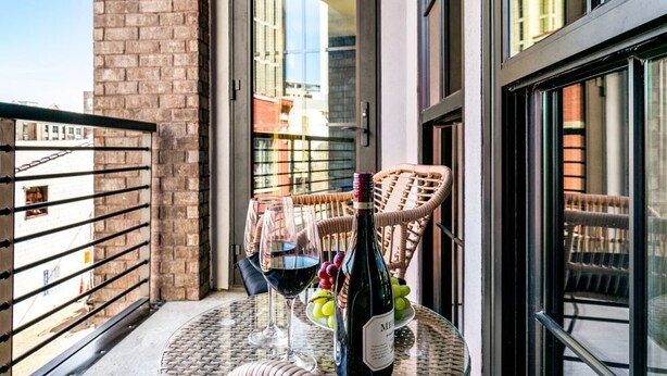 A balcony with a wicker chair, a table with a bottle of wine, glasses, and grapes.