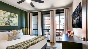 A hotel room with a bed, a desk, a ceiling fan, a large window with curtains, and a cityscape view.