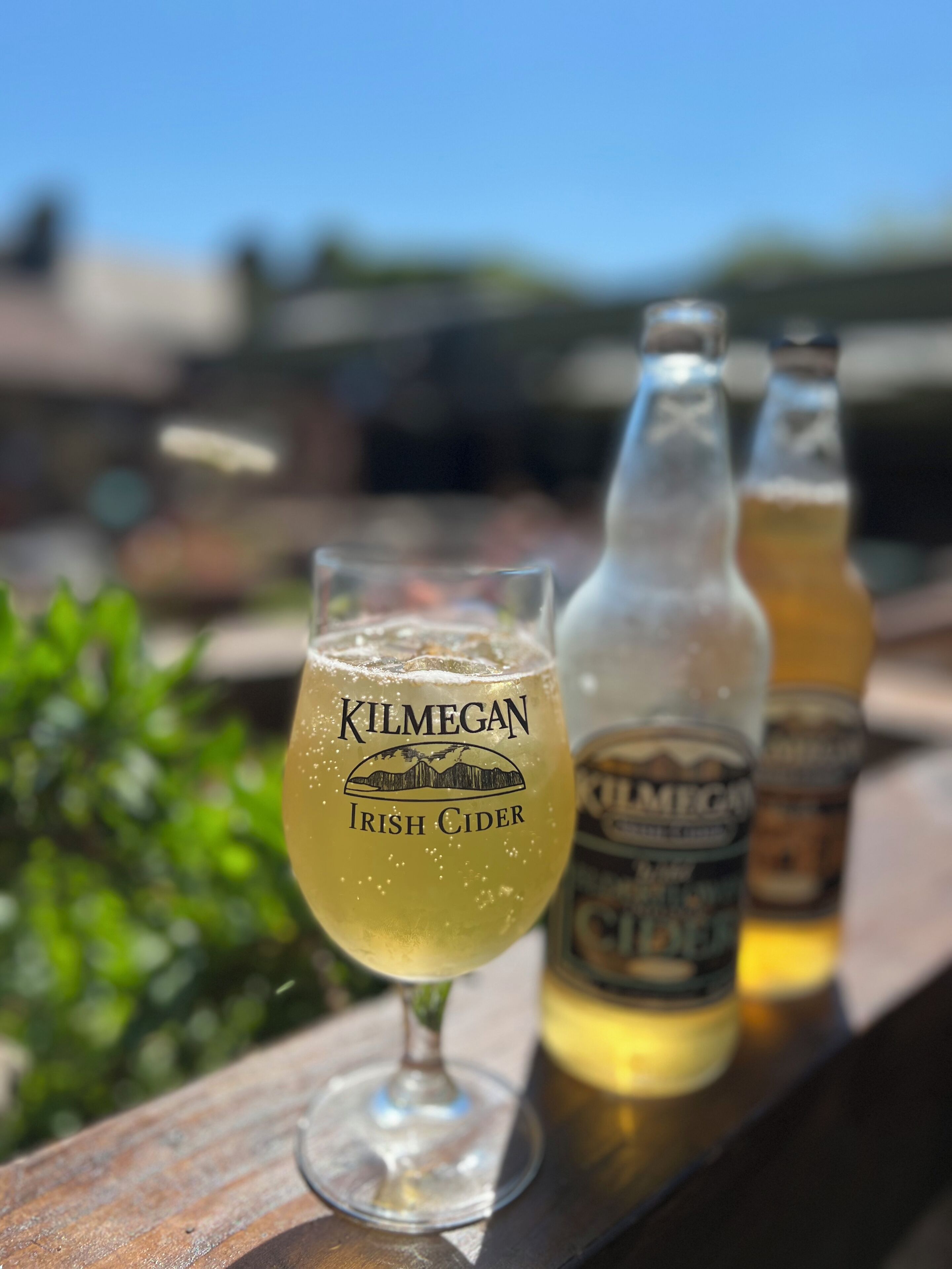A glass of Kilmegan Irish Cider on a wooden surface with two bottles of the same brand in the background.