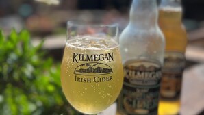 A glass of Kilmegan Irish Cider on a wooden surface with two bottles of the same brand in the background.