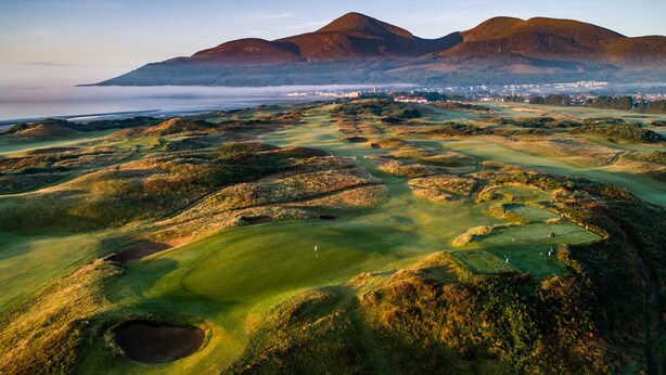 A golf course with undulating terrain and bunkers, surrounded by mountains and a coastal town in the distance. - Dundrum Inn (Newcastle)