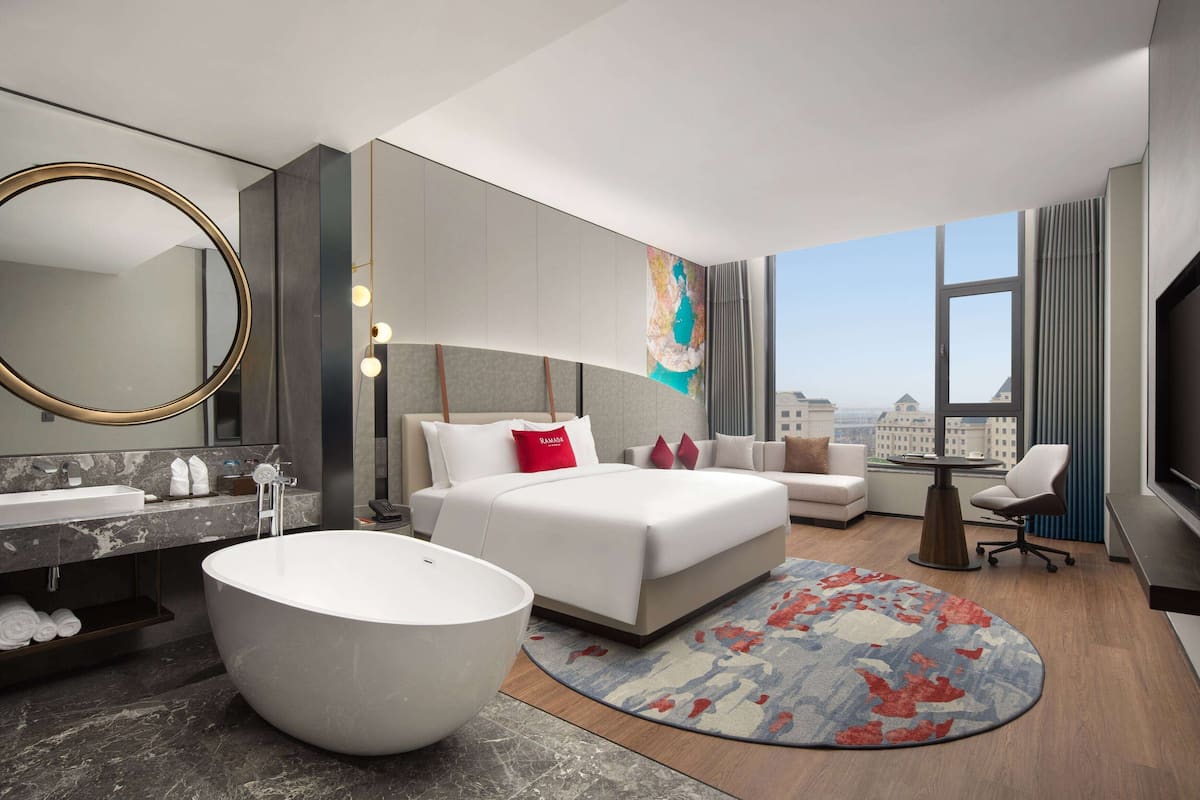 Une chambre d’hôtel moderne dotée d’un grand lit, d’une baignoire ronde, d’un coin salon et d’une grande fenêtre donnant sur la ville.