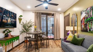 A modern living room with a dining area, a ceiling fan, a large window with curtains, and a painting of colorful buildings.