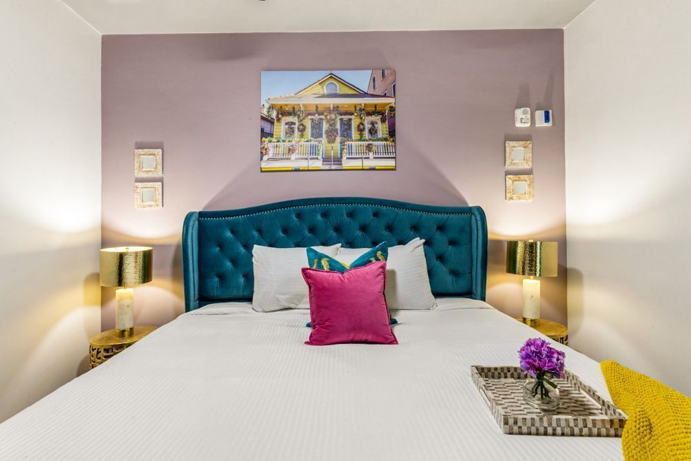 Un dormitorio con cabecera capitonada, cubrecama blanco, almohada rosa y un cuadro enmarcado en la pared.