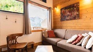 A cozy room with a wooden floor, a brown sofa with patterned cushions, a wooden chair, a small table, and a window with curtains. - 10th floor, view valley, ski locker, 36m², Valmorel (Valmorel)