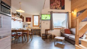 A cozy living room with a sofa, a dining area with wooden furniture, a TV, and a staircase. - 10th floor, view valley, ski locker, 36m², Valmorel (Valmorel)