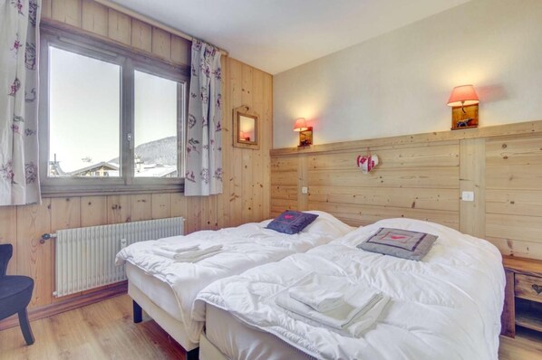 A room with two beds, a window with curtains, a radiator, and wall-mounted lamps.