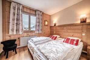 A cozy bedroom with two beds, wooden paneling, a window with curtains, a chair, and wall-mounted lamps.