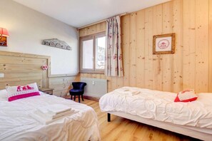 A room with two beds, a chair, a window with curtains, a radiator, and a framed picture on the wall.