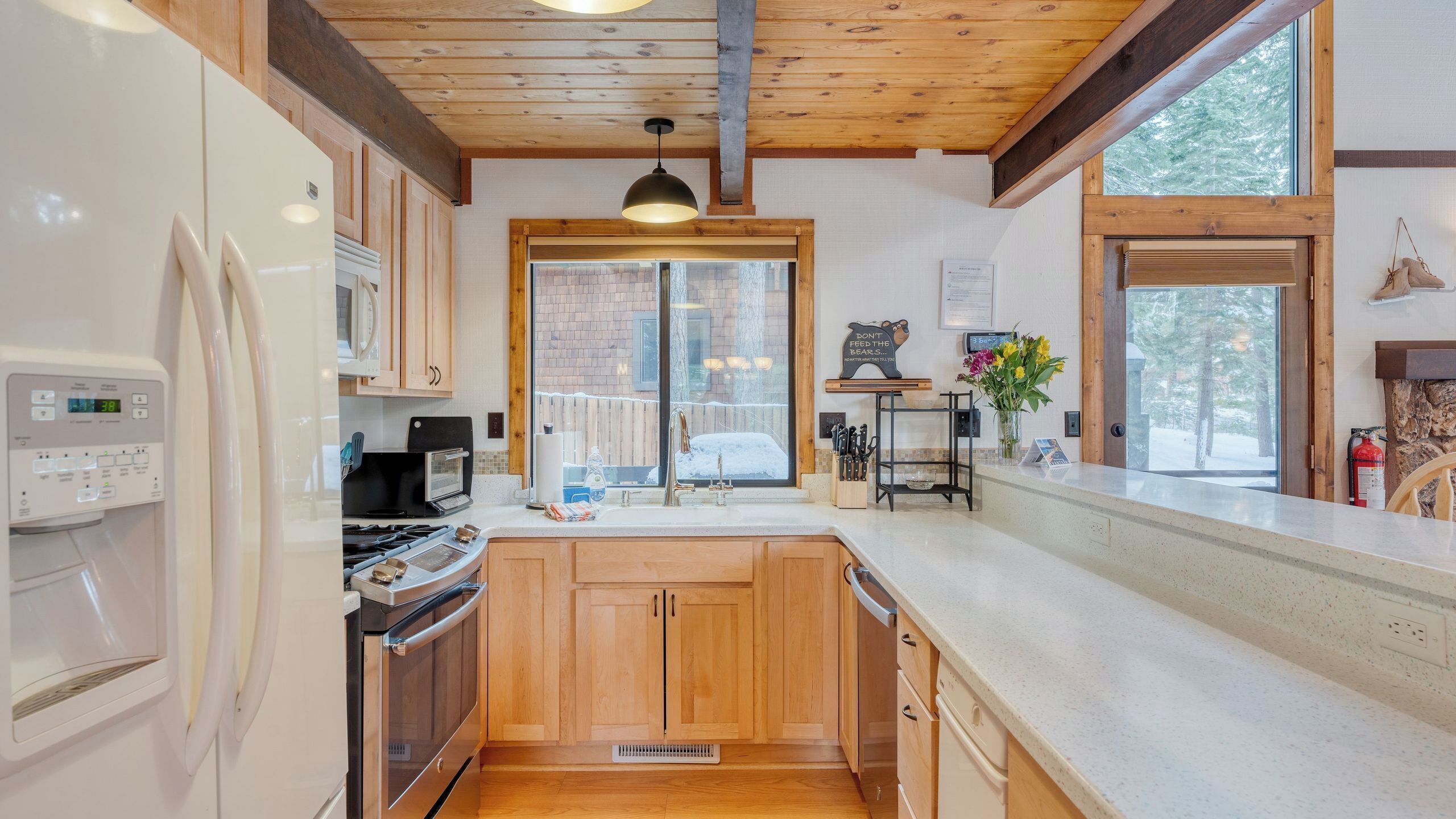 A modern kitchen with wooden cabinets, a stainless steel refrigerator, a stove, and a window with a view of a snowy landscape.