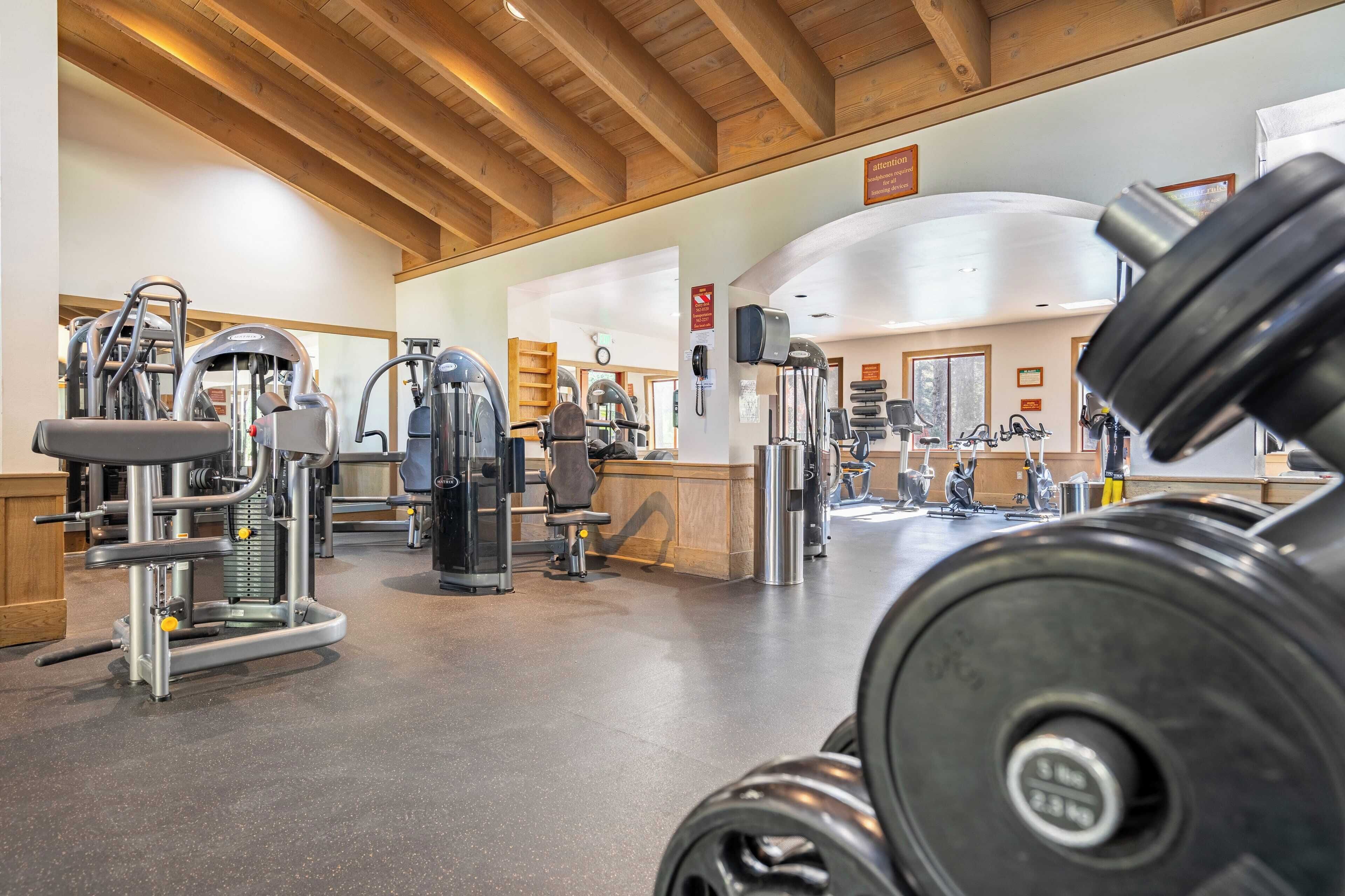 Une salle de sport équipée de divers appareils d’exercice, avec des poutres en bois au plafond et un espace d’accueil à l’arrière-plan.