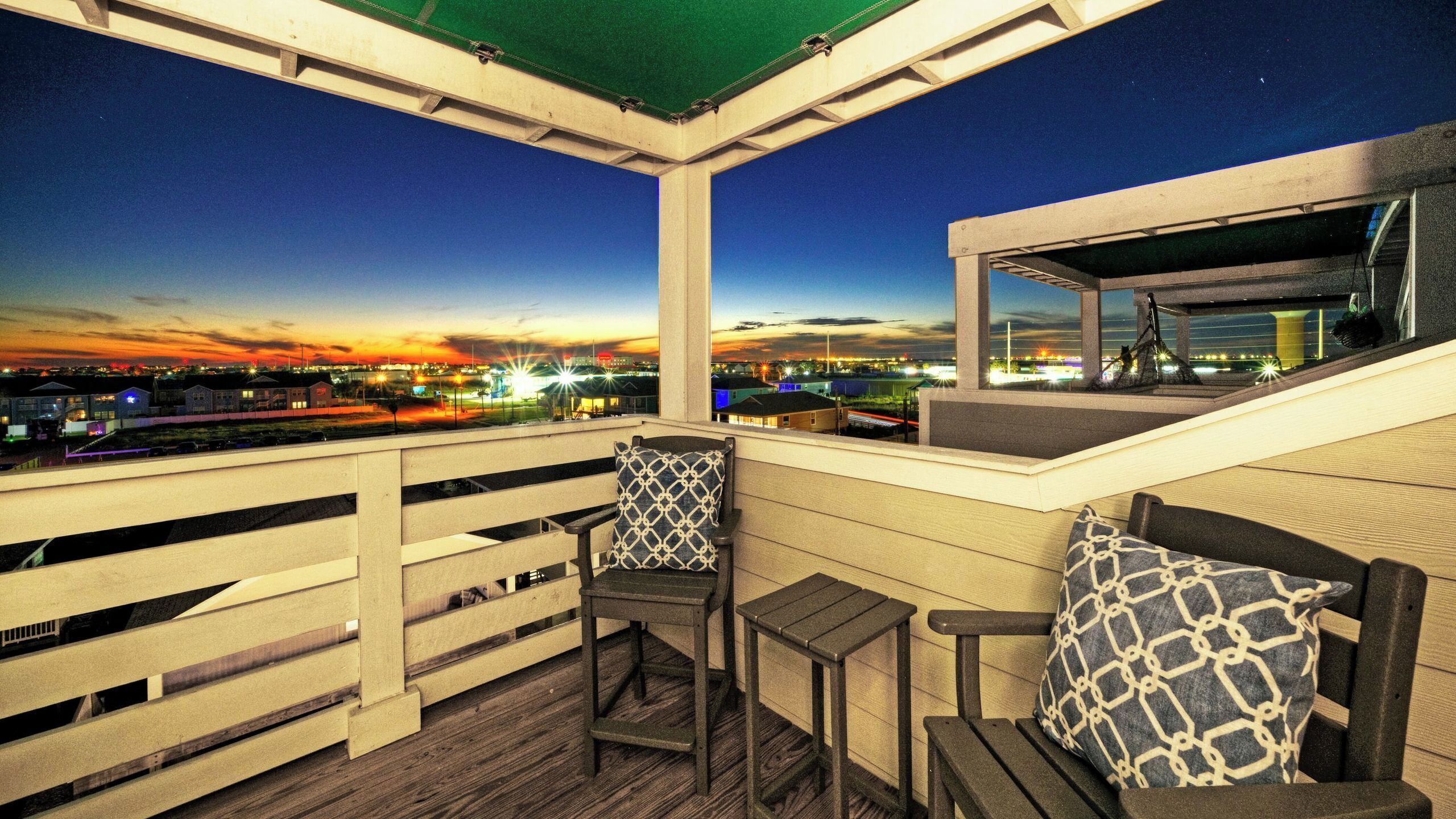 Ein Balkon mit Blick auf eine städtische Szene im Abenddämmerung, mit Holzbalkonen und zwei Stühlen mit gemusterten Kissen.