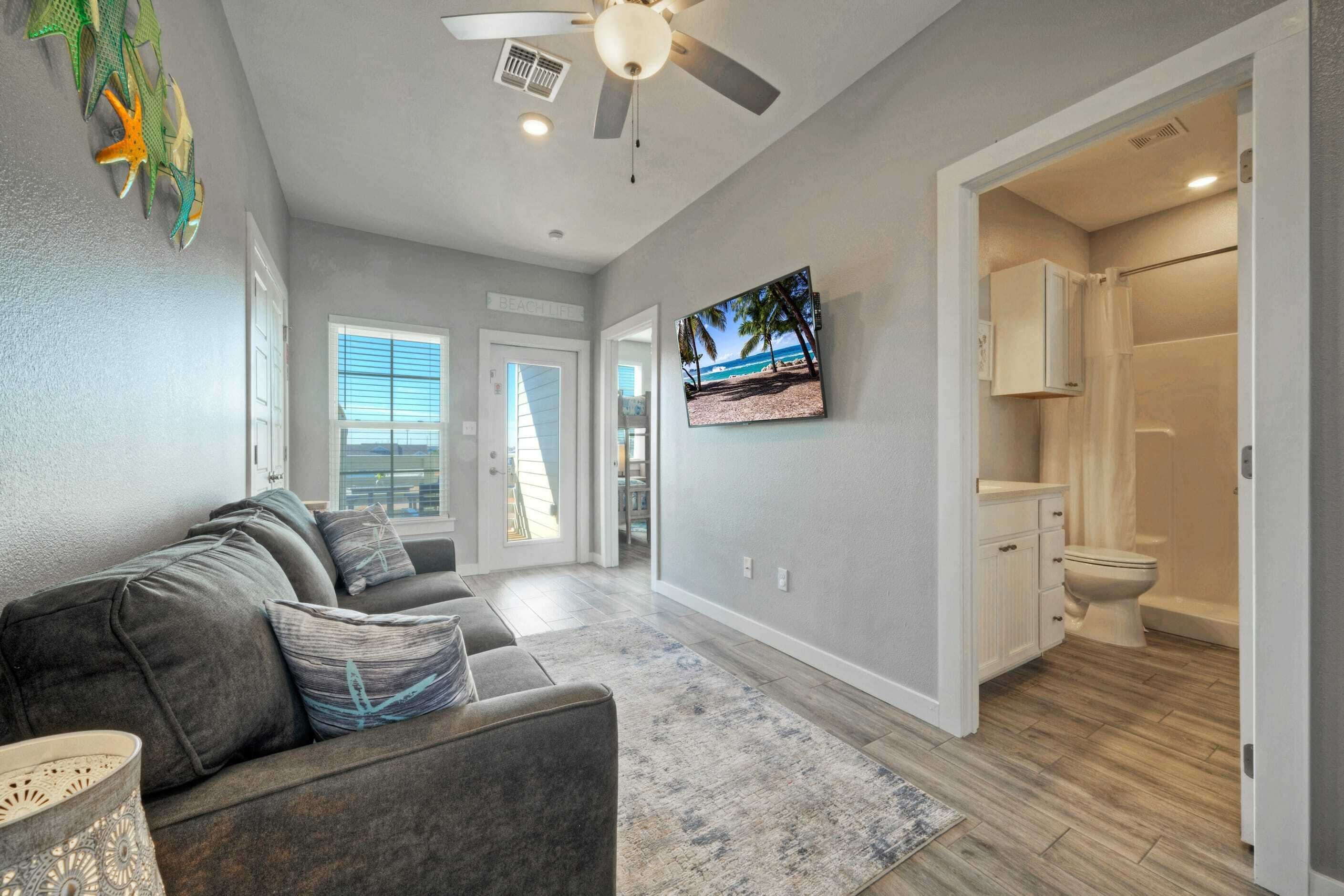 A modern living room with a gray couch, a ceiling fan, and a TV on the wall.