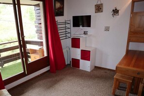 A room with a wooden desk, a television mounted on the wall, a radiator, and a window with a red curtain.