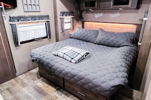 Un dormitorio con cama, cabecera, ventana con cortinas, televisor y una pequeña estantería.