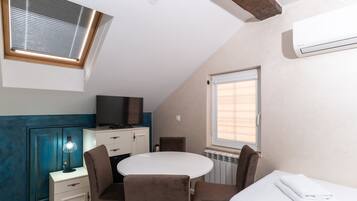 A compact room with a round table, two chairs, a bed, a TV, and a window with blinds.