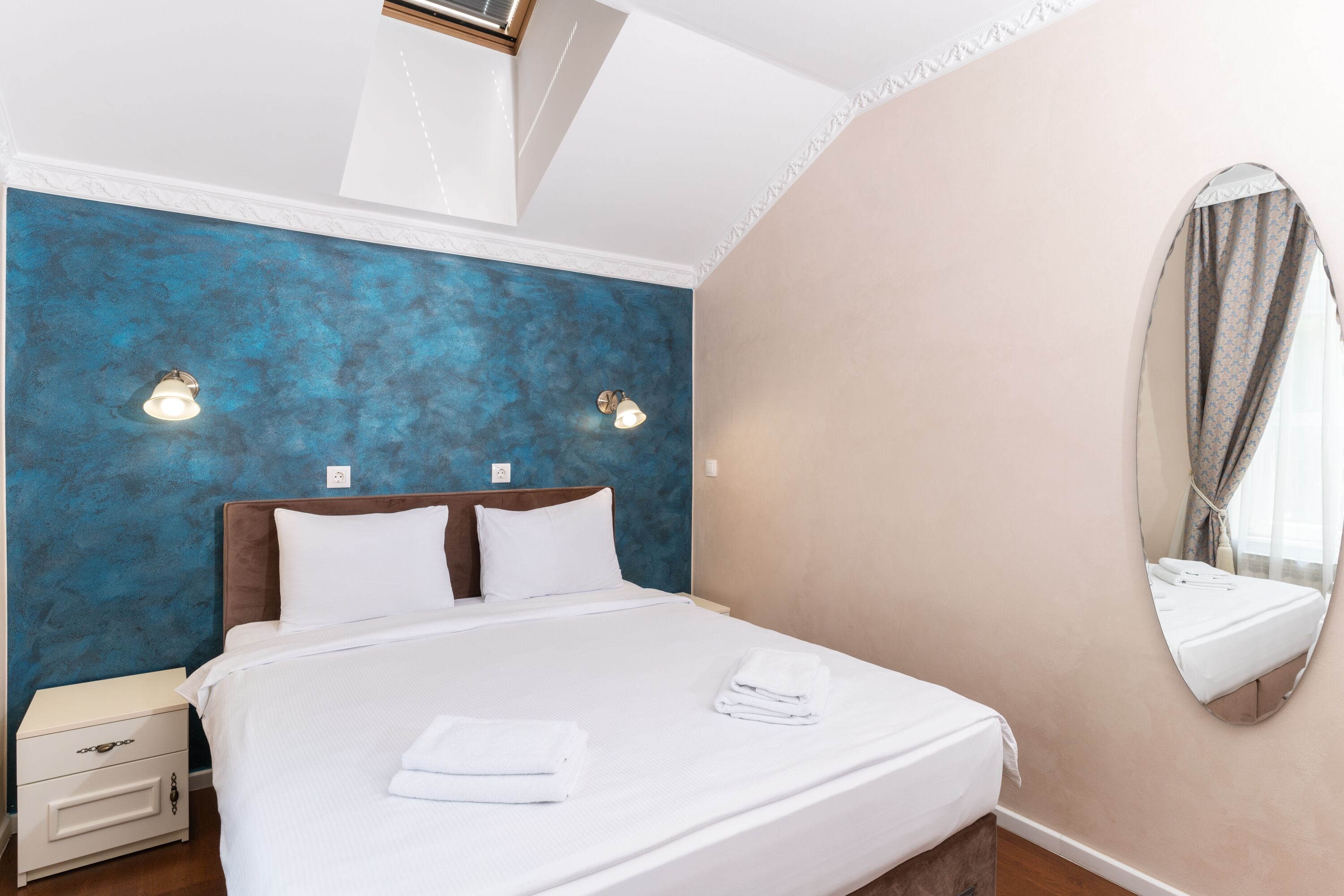 A neatly made bed with white linens and folded towels, a bedside table with a lamp, and a large mirror on the wall.