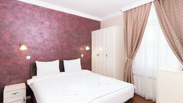Una cama bien hecha con sábanas blancas, una mesita de noche con cajón, un armario y una ventana con cortinas traslúcidas.