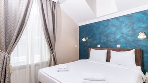 A neatly made bed with white linens and two pillows, a blue textured wall behind it, and a window with sheer curtains. - Ornament Hotel (Sarajevo)